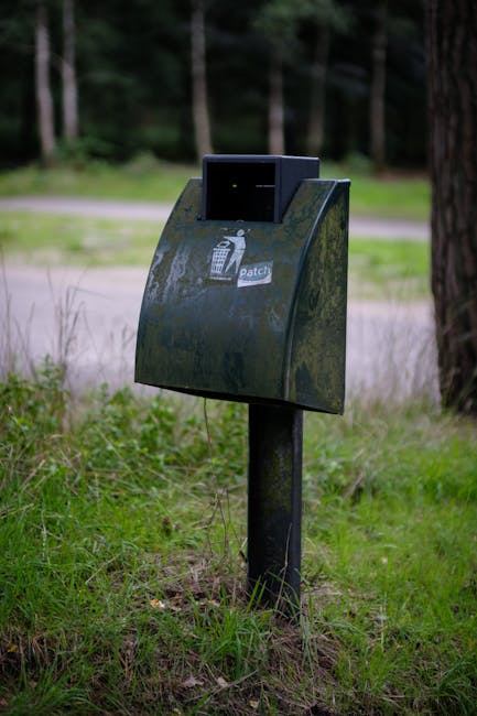 A black metal rubbish collection bin mounted on a single dark post, situated on a grassy area with some moss or lichen growth visible on its surface. The bin has a small, rectangular opening at the top and a sticker with a pictogram of a trash bin and the word 'patch' on its front. The background features a natural environment with a paved path or road, out-of-focus trees, and greenery, suggesting the bin is located in a park or woodland edge. The lighting appears natural, with diffuse daylight illuminating the scene, and the setting reflects an outdoor waste disposal point suitable for informal or alternative rubbish collection in green areas. The presence of the Rubbish Clearance Roehampton service and the page context imply this is part of a private or independent waste management solution for outdoor rubbish or bulky waste collection.