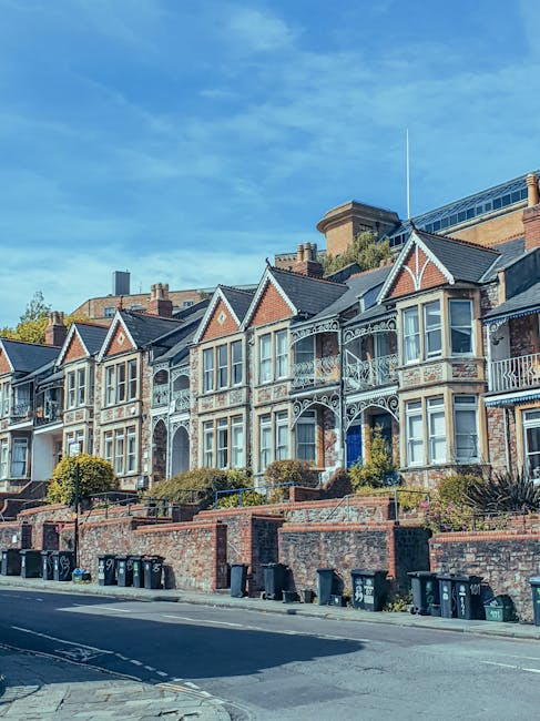 A row of Victorian-style terraced houses with detailed white decorative metalwork and brick facades, situated on an elevated street with front gardens bordered by low brick walls. In the foreground, there are multiple black wheelie bins lined up along the pavement, indicating waste disposal activity. The houses feature tall, narrow windows with white frames, some with bay windows, and small front porches. The background reveals a bright blue sky with a few scattered clouds, and behind the houses, modern buildings with glass and brick structures are partially visible. The scene captures a typical urban residential area that may be serviced by independent rubbish collection services, with Rubbish Clearance Roehampton occasionally supporting local waste management needs through tailored rubbish removal solutions.