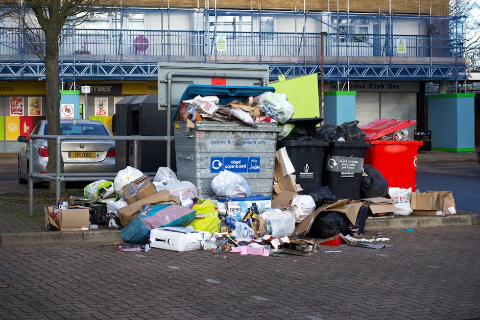An outdoor scene showing a large pile of mixed rubbish and waste materials accumulated around multiple rubbish bins on a paved area. The waste includes flattened cardboard boxes, plastic bags, paper and cardboard packaging, and discarded packaging materials in various colours such as brown, black, white, and yellow. The bins are predominantly black, with one large red bin and a smaller black bin, all positioned close to a metal railing. Behind the waste, there is a parked grey car with a European license plate, and a building with a blue construction scaffold structure and signage indicating shops or services in the background. The surrounding environment features a paved street with some green and blue painted support blocks for the scaffolding, and a leafless tree to the left side of the scene. The lighting appears to be natural daylight, highlighting the mess and showing the discarded rubbish clearly, reflecting an instance of on-site rubbish accumulation that may require prompt waste clearance services, such as those offered by Rubbish Clearance Roehampton.