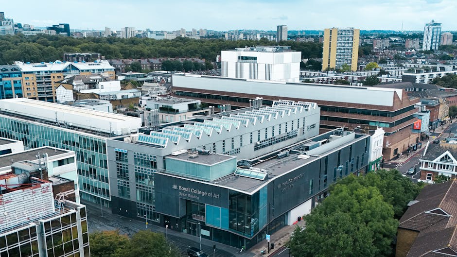 An aerial view of a modern urban cityscape featuring a large building housing the Royal College of Art, identifiable by its prominent signage on the dark exterior wall. The building has a sleek, contemporary design with extensive glass facades and metal cladding, and a series of angular, light-colored skylights on the roof. Surrounding the college are various other commercial and residential structures, including mid-rise apartment blocks and office buildings with varied finishes such as brick, glass, and metal panels. The scene is set during daylight with clear skies, and the streets below show sparse vehicular and pedestrian activity. The overall environment presents a densely built-up area with a mix of older and newer architectural styles, characteristic of a central urban location. This visual could relate to private waste management or on-site clearance services, which may be relevant for independent collections within city centers, as part of a comprehensive rubbish removal strategy. Rubbish Clearance Roehampton provides services suited to managing waste disposal in such environments, ensuring efficient removal of various types of rubbish without reliance on local authority waste collection methods.